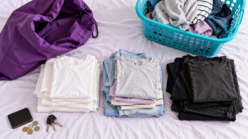 Roupas separadas em pilhas de brancas, claras e escuras sobre a cama, com sacola, cesto, carteira e chaves